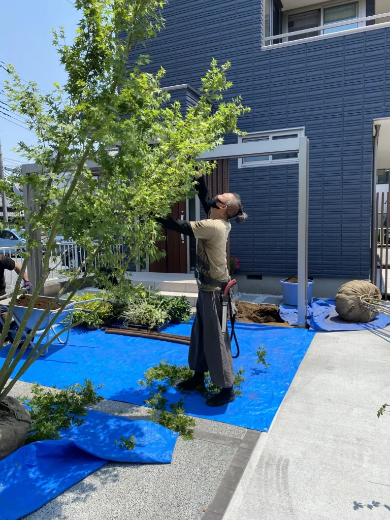 植栽の施工中の様子。奥にLIXILプラスGのアーチフレームと建物ファサードが見え、雑木系の植栽がこれから外構全体に彩りを添えていく場面。