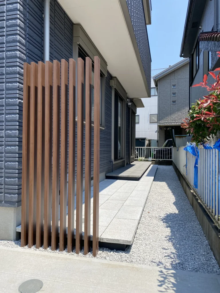 建物側面に設置された木調アルミ縦格子のアップ。目隠し効果を持ちながら外構全体のデザイン統一感を担い、横には大判タイルを敷いた浮き階段風の広々としたデッキと白砂利が続くローメンテナンス仕上げ。